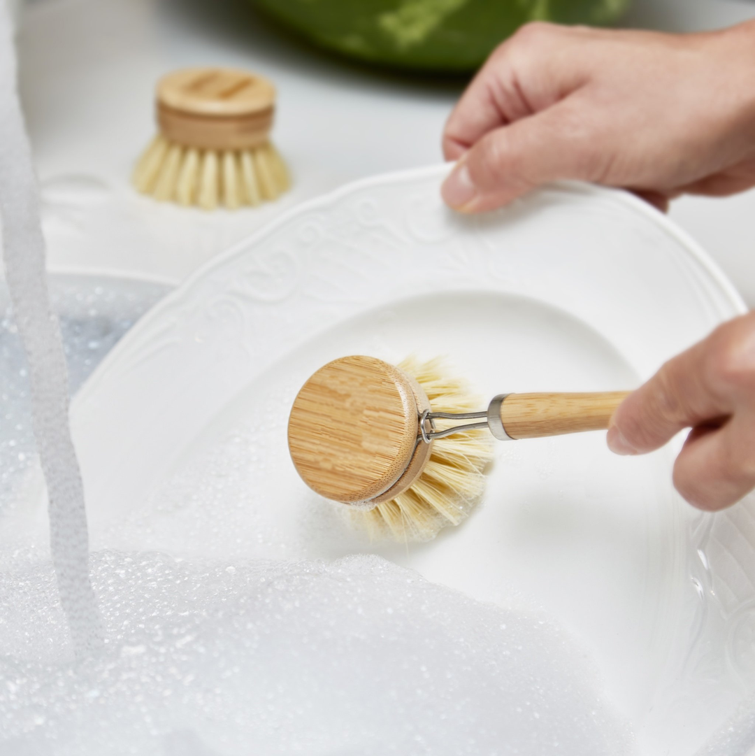 Person using a wooden dish brush to clean a white plate with soap suds.